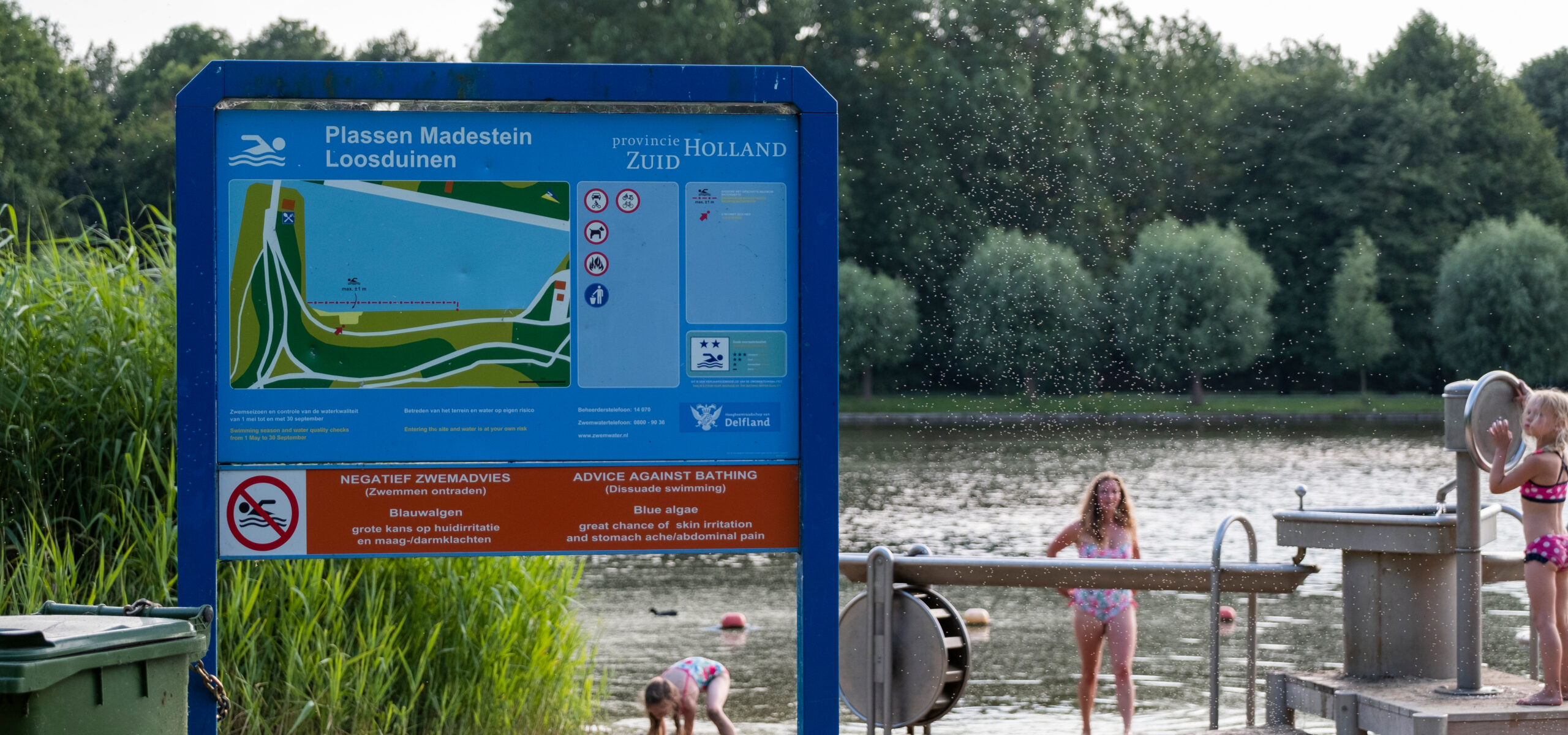 Een bord in de openbare ruimte waarop gewaarschuwd wordt voor blauwalg. Op de achtergrond een strandje en zwemmeer met een moeder en een kind in badkleding.