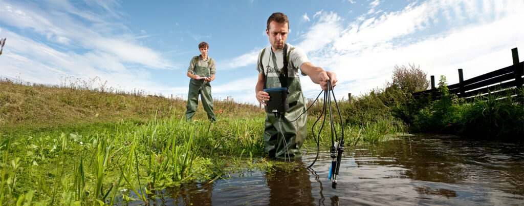 Twee mensen in waadpakken voeren metingen uit in slootwater van een ondiepe boerensloot in een landelijke omgeving. Een persoon staat met meetapparatuur in het water en dat ander op de kant op meting te schriftelijk te noteren.