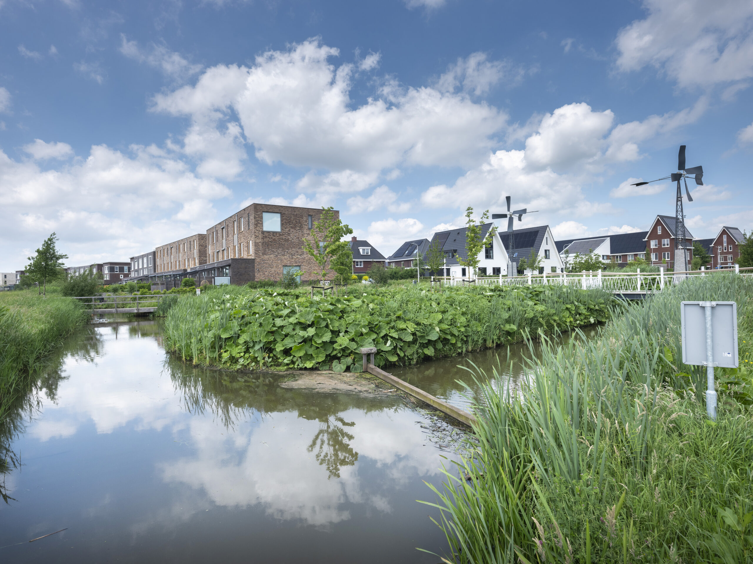 Nederlands landschap met waterweg, groene vegetatie, moderne huizen en een windmolen onder een blauwe lucht