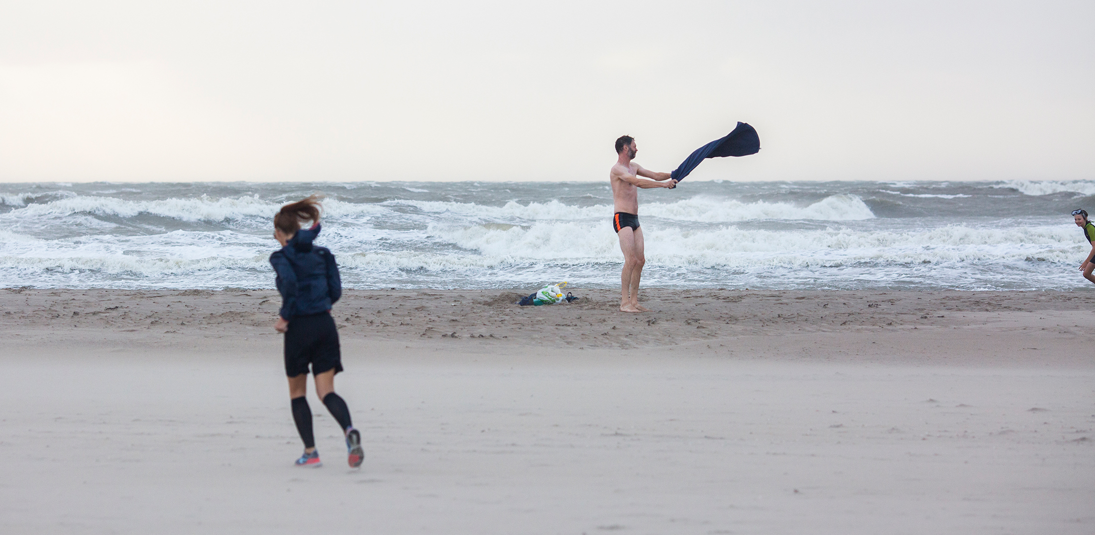 Stormachtig weer aan de kust. Zand verwaait aan het strand, ruwe golven. Een man in zwembroek heeft een handdoek vast die meewaait met de wind. Op de voorgrond nog een vrouw die aan het joggen is.