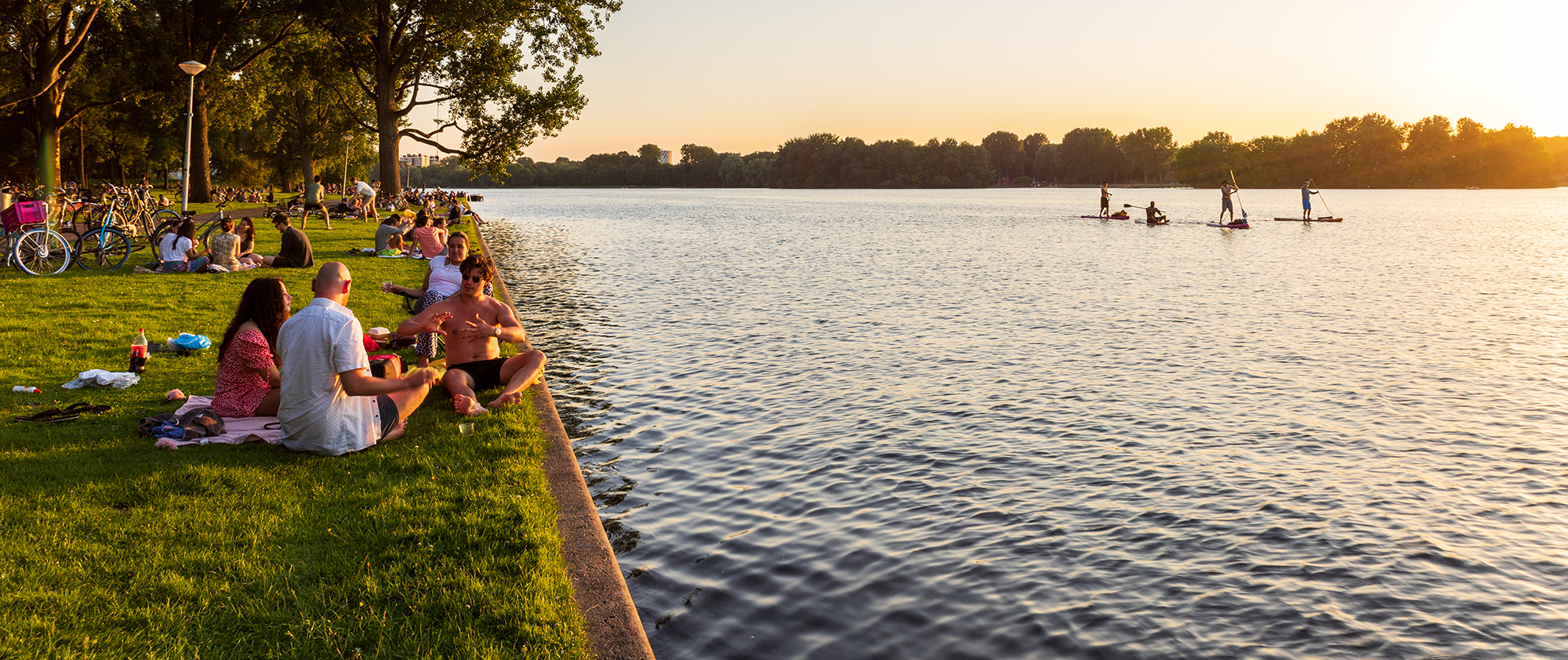Recreatie gebied met water en grasveld. Er zijn suppers op het water en op het grasveld zitten jongeren te chillen bij zonsondergang.