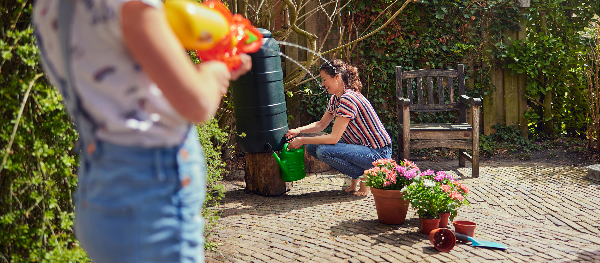Een vrouw met een gieter die water uit haar regenton wil tappen in de tuin. Zij wordt onderstussen na gesproten door een meisje met een oranje waterpistool.