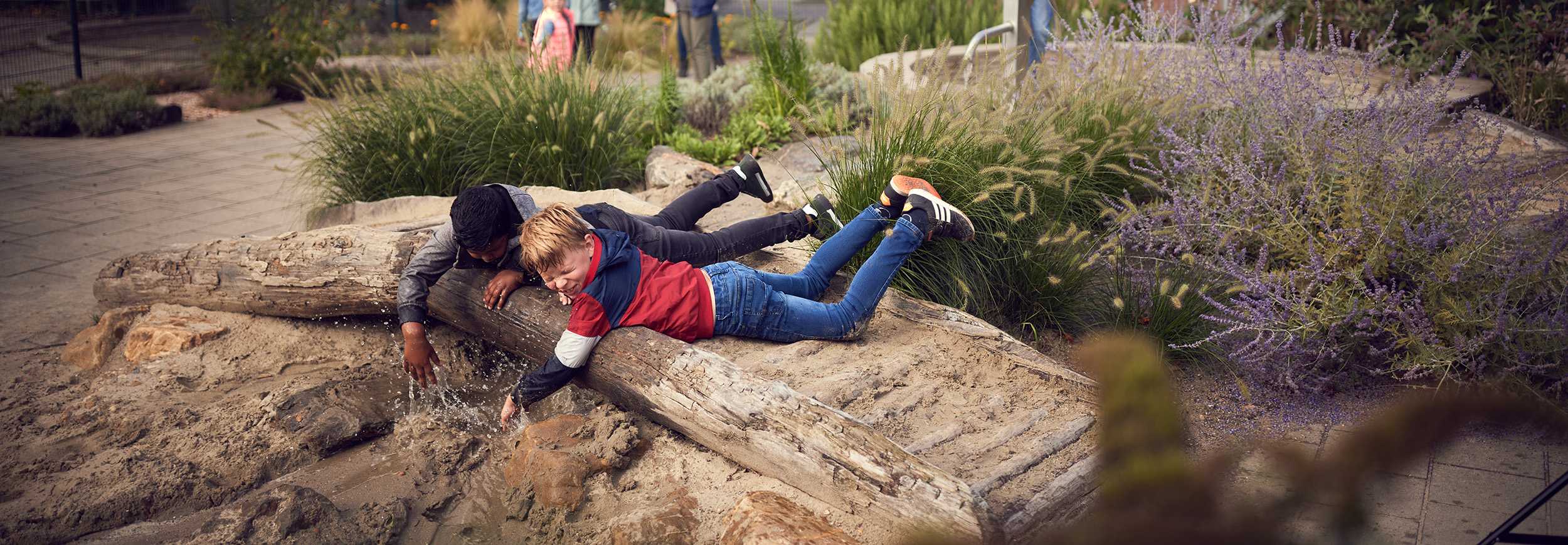 2 Jongens van ongeveer 8 jaar oud liggen op een houte bruggen dat over een wadi ligt met zand er in. Ze zijn met het water aan het spelen dat onder de brug door de wadi loopt.