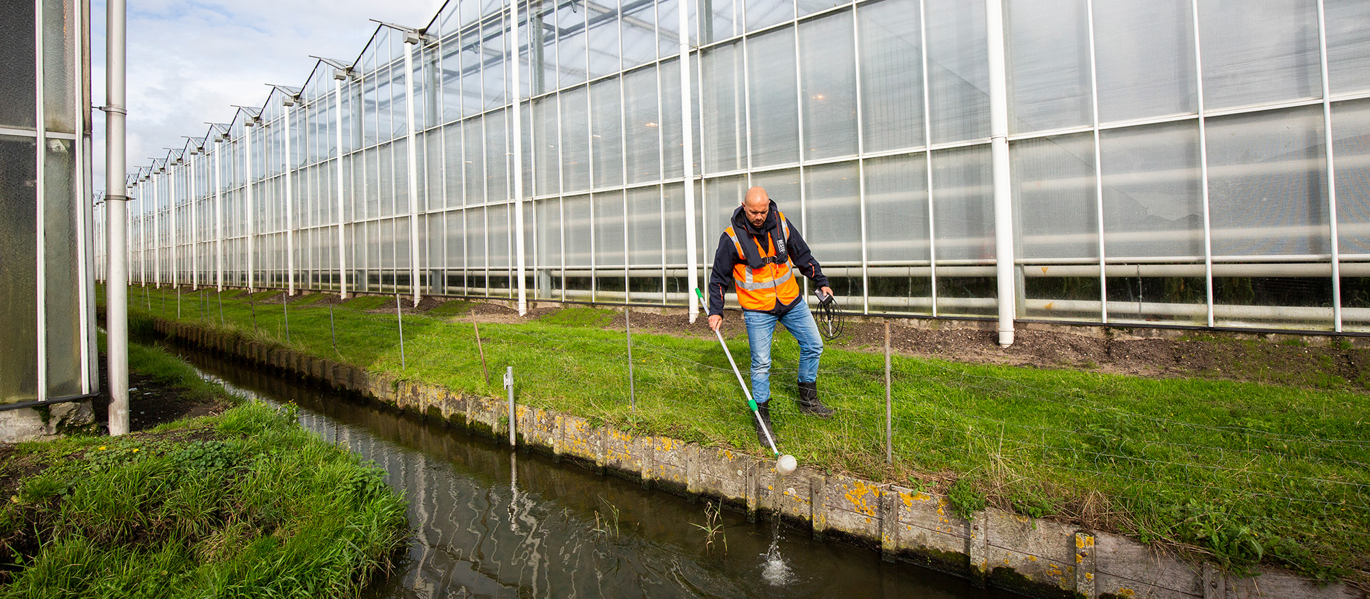 Waterschapper neemt monster van sloot water. De sloot ligt tussen kassen.