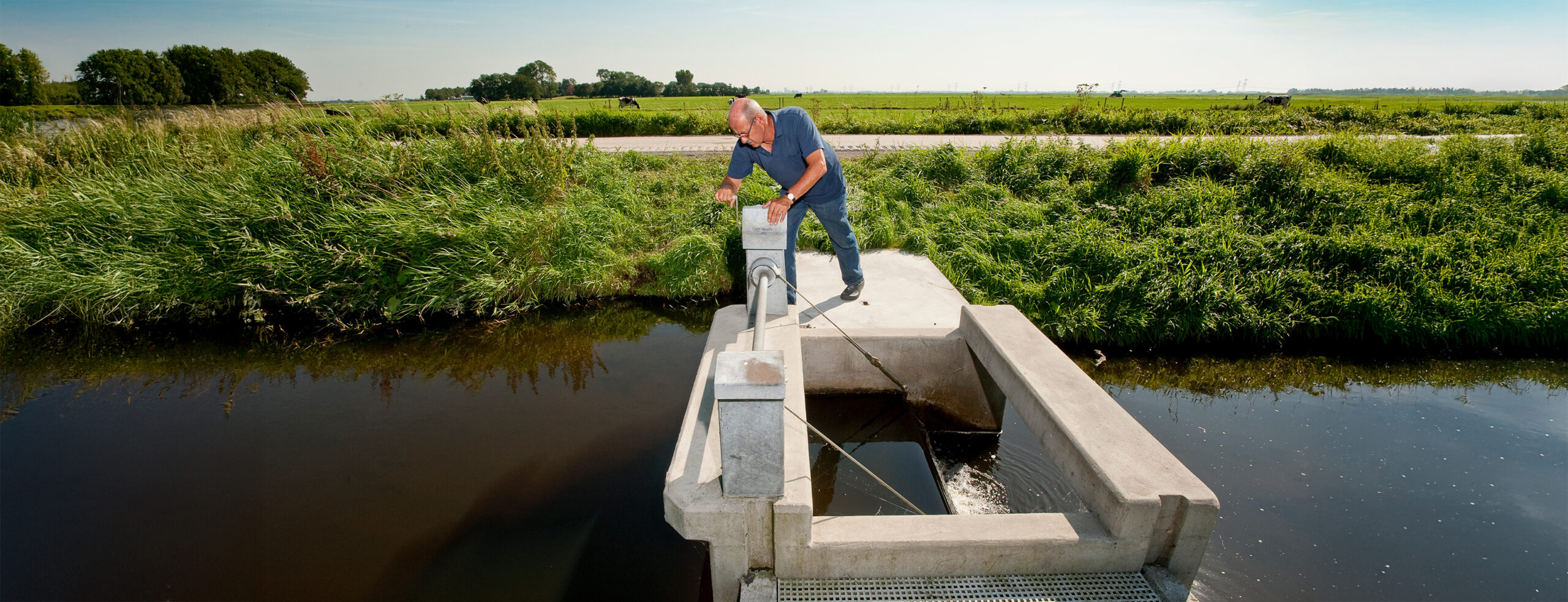 Een polderlandschap met op de voorgrond water met daarin een stuw. Op de stuw staat een waterschapper om de stuw te bedienen.