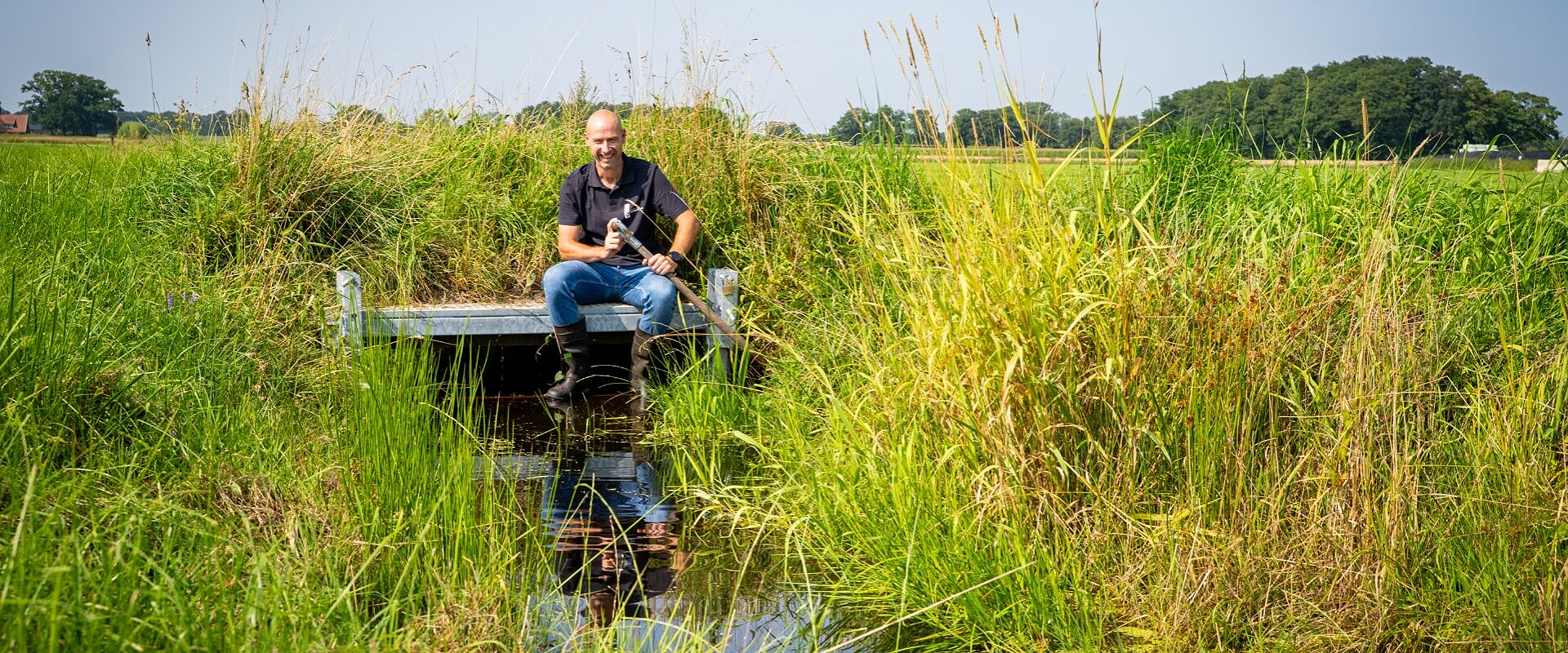 Waterschapper zit op stuw in landelijke gebied bij een klein slootje waar nog water instaat. De lucht is strak blauw.