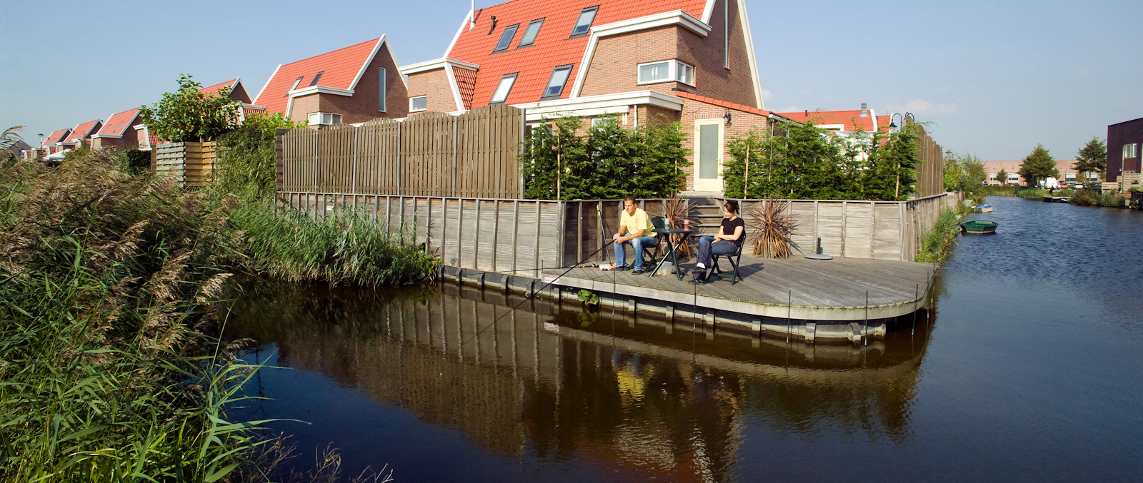 Woning met steiger aan het water. Op de steiger zitten een man en een vrouw. De man is aan het vissen.