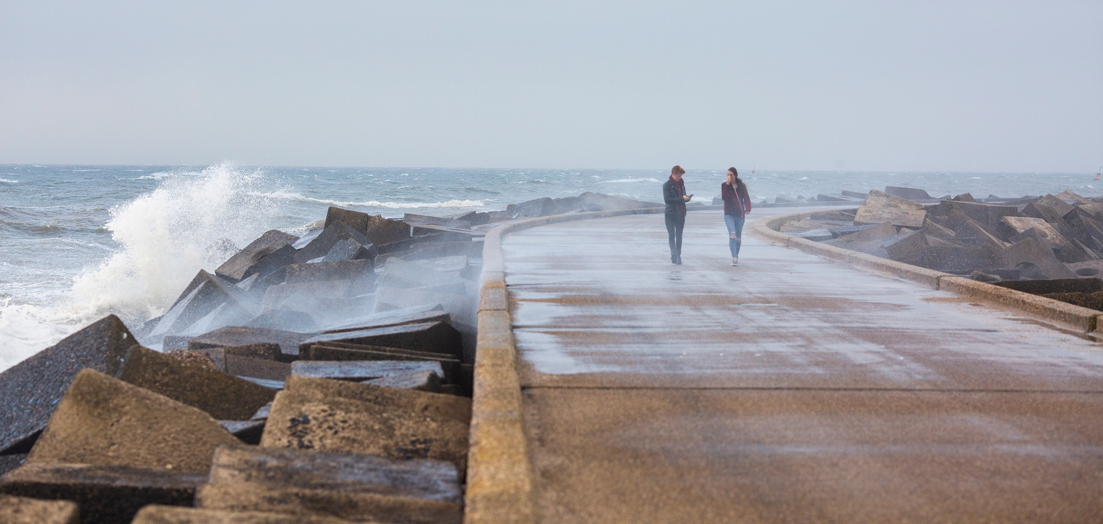 twee personen lopen over kade met storm, water slaat over de kade heen.