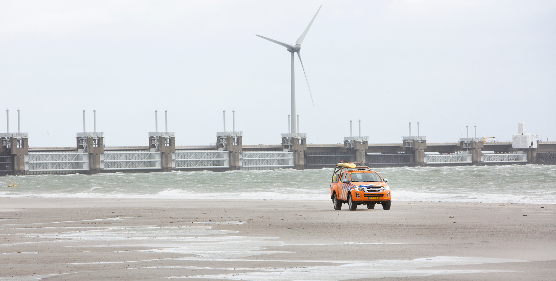 Stormachtig weer. Auto Rijkswaterstaat op het strand met op de achtergrond de Zeelandbrug. Onderdeel Deltawerken.