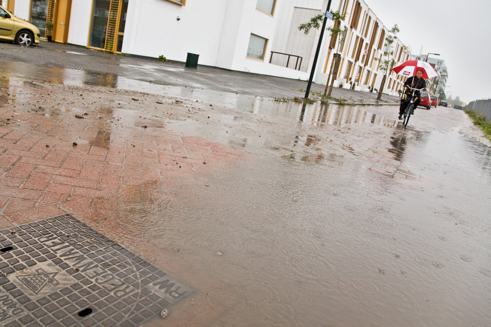 Wateroverlast doordat als gevolg van hevig regen plassen op straat blijven staan. Fietser met paraplu rijdt door de plassen.