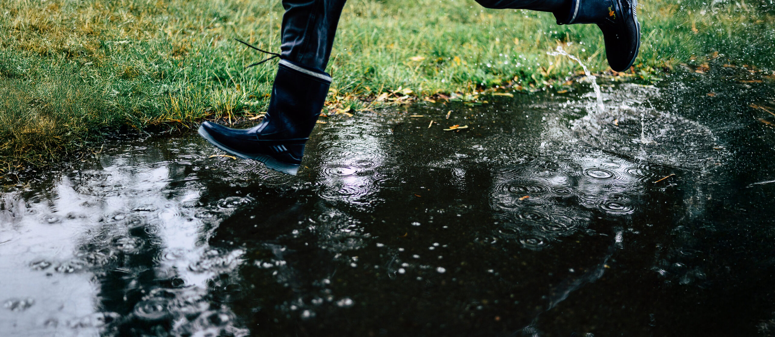 Close-up beeld van de straat met gras erlangs waarop het regent. je ziet een paar blauwe laarzen die door de regen rent.