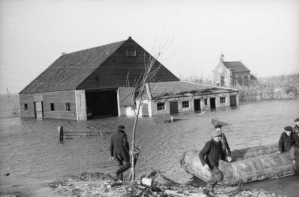 Watersnoodramp 1953, oude zwart-wit foto: Boerderij deels onder water. Op de voorgrond mannen in een rubberboot.